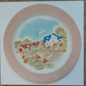 May include: A decorative ceramic plate featuring a hand-painted rural landscape scene. The design includes houses with red and blue roofs, a road, and a blue sky. The plate has a light pink border and is signed "Aimm".