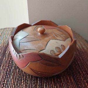 May include: A wooden bowl with a lid, featuring a carved design of stylized leaves and flowers in shades of brown, red, and white. The bowl has a rounded shape and a slightly flared rim.
