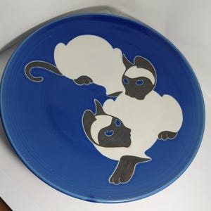 May include: A round, blue ceramic plate featuring a Siamese cat design. The cats are depicted in white with dark brown accents, including their faces, ears, and paws. The plate's rim is a darker shade of blue.