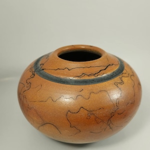 Könnte beinhalten: Eine runde, braune Keramikvase mit einem dunkelblauen Band in der Mitte. Die Vase hat einen schwarzen Innenraum und ist mit schwarzen, wellenförmigen Linien auf der Oberfläche verziert. Die Vase hat eine glatte, matte Oberfläche.