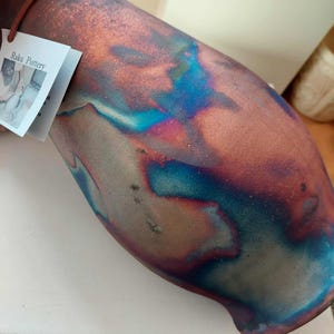 May include: A ceramic vase with a metallic finish featuring swirling iridescent colours of copper, bronze, blue, and purple. A white paper tag is attached to the neck of the vase, displaying the text Raku Pottery and a small photograph.