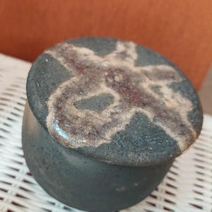 May include: A small, round, dark grey ceramic jar with a matching lid. The lid features a textured, irregular cross-shaped pattern in a lighter, brownish-white glaze that contrasts with the matte, dark grey surface of the container body.