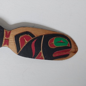 Puede incluir: Talla de pez de madera con un diseño detallado. El pez es principalmente de color madera natural, con detalles pintados en negro, rojo y verde. La talla presenta formas y líneas estilizadas, mostrando un estilo de arte tradicional.