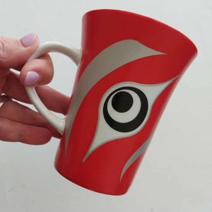 May include: Red ceramic mug with a white handle. The mug features a stylized design in silver and black, resembling an eye. The mug is held by a hand with pink nail polish.