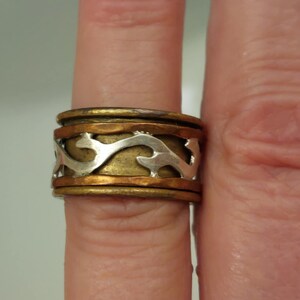 May include: A mixed-metal ring with a silver, gold, and copper design. The ring features a silver, wavy pattern set against a gold band, with copper accents. The ring is on a finger.