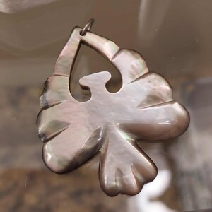 May include: A carved mother-of-pearl pendant shaped like a stylised bird in flight. The iridescent shell material displays shades of grey, brown, and silver with a smooth, polished surface. A small metal jump ring is attached to the top point.