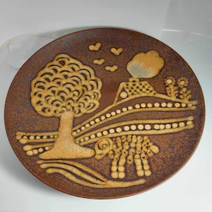 May include: A brown ceramic plate with a stylized design of a tree, a house, and a flower. The design is in shades of brown and yellow.