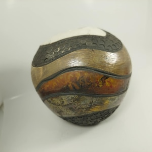 Könnte beinhalten: Eine kugelförmige Keramikvase mit strukturierter Oberfläche. Die Vase hat horizontale Farbstreifen, darunter Weiß, Schwarz, Braun und Orange. Das schwarze Band hat ein gepunktetes Muster. Die Vase hat ein glänzendes Finish.