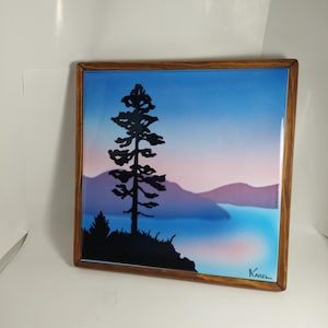 Pode incluir: Obra de arte quadrada emoldurada com uma paisagem serena. Uma silhueta preta de uma árvore se destaca contra um lago e montanhas, com um céu gradiente de azul, rosa e roxo. A moldura é de madeira.