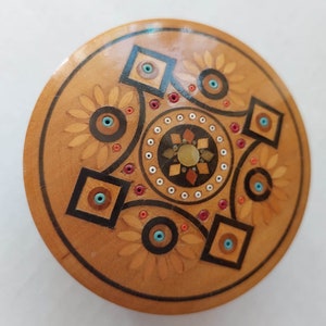 Peut inclure: Une boîte ronde en bois avec un décor en incrustation. Le motif présente un cercle central avec un motif en étoile, entouré de quatre carrés avec des accents bleus et blancs. La boîte est en bois clair avec une finition naturelle.
