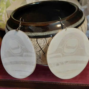 May include: A pair of oval-shaped earrings featuring a light cream-colored surface with a subtle, etched graphic design of a stylized animal form. The earrings are suspended from silver-toned metal hooks and have a smooth, matte finish.