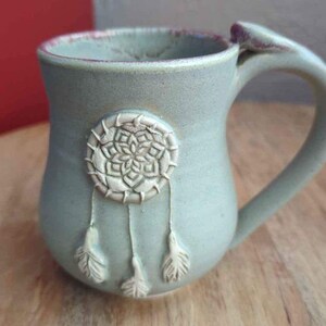 Pode incluir: Uma caneca de cerâmica verde claro com um design de apanhador de sonhos branco e penas. A caneca tem um corpo arredondado e uma alça.