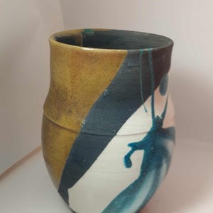 Könnte beinhalten: Eine handgefertigte Keramikvase mit einem einzigartigen Design. Die Vase zeigt eine Kombination aus Farben, darunter Gelb, Dunkelblau, Weiß und Türkis. Das Design ist abstrakt, mit einem diagonalen gelben Abschnitt und einem dunkelblauen Abschnitt. Türkisfarbene Farbe tropft auf den weißen Abschnitt.