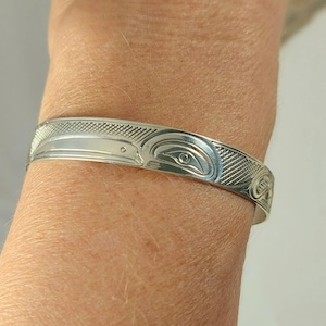 Może przedstawiać: Srebrna bransoletka z mankietem z szczegółowym wzorem rdzennych Amerykanów. Bransoletka posiada grawerowany motyw ptaka lub zwierzęcia, ze skomplikowanymi wzorami i teksturami. Bransoletka jest noszona na nadgarstku.