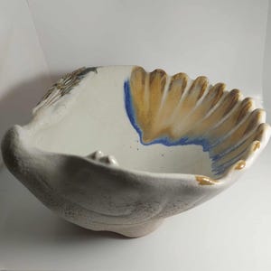 Pode incluir: Uma tigela de cerâmica artesanal com um design de concha. A tigela é branca com detalhes dourados e azuis. A borda tem uma borda ondulada. Uma peça de arte única.