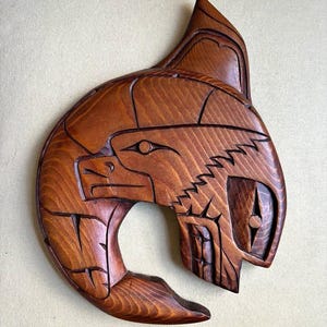 May include: A hand-carved wooden wall plaque featuring a stylized eagle head design. The medium-brown wood displays a visible grain pattern with deep, precise grooves forming the bird's eye, beak, and feathers in a curved, crescent-like shape.