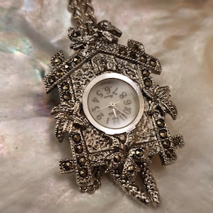 May include: A silver-toned pendant watch in the shape of a cuckoo clock. The watch face is white with black numerals and hands. The clock's decorative frame is detailed with small, dark accents and a chain.