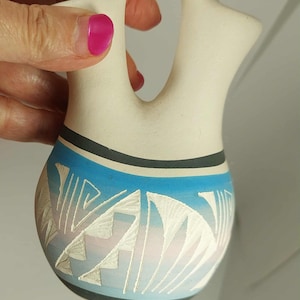 Puede incluir: Un jarrón de cerámica color crema con un diseño de asa único. El jarrón presenta un patrón geométrico azul y blanco alrededor de la base, con bandas negras y grises. El jarrón es sostenido por una persona con esmalte de uñas rosa.