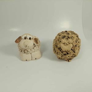 May include: Two ceramic sheep figurines. One is off-white with brown accents on the ears and a detailed face. The other is a textured, brown sheep with a smiling face. Both are on a white background.