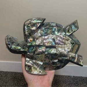 Puede incluir: Una figura decorativa de tortuga con un caparazón de mosaico. El caparazón está hecho de piezas iridiscentes de concha de abulón en tonos verdes, azules y morados. La figura tiene una base y una cabeza oscuras.
