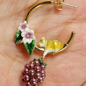 May include: Gold-tone hoop earring with a squirrel, flowers, and a cluster of purple beads. The squirrel is yellow and white, perched on the hoop. The flowers are white and pink with green leaves. The cluster of beads is purple.