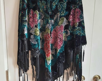 May include: A black velvet shawl featuring a vibrant floral pattern in shades of pink, green, and blue. The garment is adorned with intricate swirling designs and small pink accents, finished with long black fringe trim along the bottom edges.