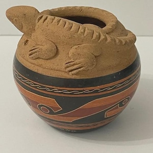 Puede incluir: Una olla de cerámica de color terracota con un lagarto esculpido en el lateral. La olla tiene una banda negra con diseños geométricos naranjas y blancos. El lagarto tiene garras y escamas detalladas. La olla es redonda con una abertura ancha.