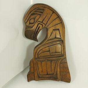 May include: A carved wooden sculpture of an eagle's head and upper body. The sculpture is a warm brown color with detailed lines and geometric shapes. The eagle's beak is curved, and the eye is visible. The sculpture is likely a decorative item or wall hanging.