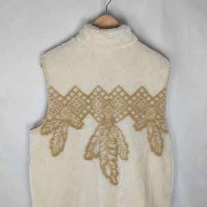 May include: A sleeveless cream-colored vest made of a soft, fuzzy material. The upper back features a tan-colored embroidered pattern consisting of a geometric diamond border above three downward-pointing leaf-like motifs.