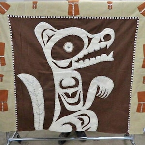 Puede incluir: Un tapiz marrón y blanco con un diseño de animal estilizado. El animal es blanco con acentos marrones y está rodeado por un borde de cuentas blancas.