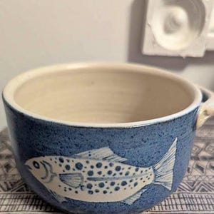 May include: A ceramic bowl with a blue exterior and a cream-coloured interior. The bowl features a white fish design with dark blue spots. The bowl has a handle and is on a patterned surface.