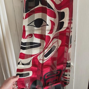 May include: A rectangular scarf featuring a bold red, black, and cream-coloured graphic design with stylised facial features and abstract shapes. The fabric has a smooth, semi-sheer finish and includes a signature in the bottom left corner.