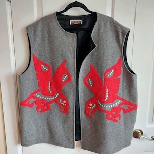 May include: A sleeveless grey vest featuring a round neckline and a black interior lining. The front displays two large, symmetrical red bird-shaped appliqués decorated with small white circular accents. A label is visible at the inner neckline.