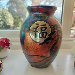 Könnte beinhalten: Eine dekorative Keramikvase mit Metallic-Finish in Rot-, Blau- und Bronzetönen. Die Vase hat ein goldenes Emblem mit asiatischen Schriftzeichen und ein schwarzes Baumdesign. Die Vase hat einen runden Körper und einen schmalen Hals.