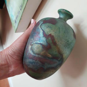 May include: A small, rounded ceramic vase with a narrow neck and a flared rim. The surface features a mottled, textured finish with blended shades of deep forest green, metallic bronze, dark brown, and hints of iridescent blue throughout the body.