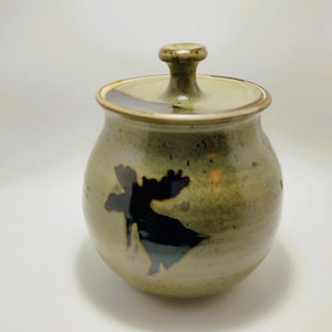 Könnte beinhalten: Eine Keramikdose mit Deckel, die eine schwarze Elch-Silhouette auf einem grün-braunen Hintergrund zeigt.