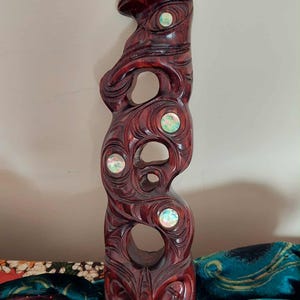 May include: A tall, dark brown wooden sculpture featuring intricate carved patterns and four circular iridescent abalone shell inlays. The vertical piece has a smooth, polished finish with several open cutouts throughout its elongated form.