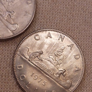 May include: Two silver Canadian dollar coins. One coin is partially visible, the other is in full view. The coin shows a canoe with figures, a tree, and the word "CANADA" and "DOLLAR". The year "1975" is also visible.