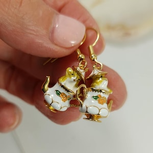 May include: A pair of gold-toned teapot earrings. Each earring features a white teapot with yellow accents, a small orange flower design, and a gold-colored handle and spout. The earrings are held by a person's hand.