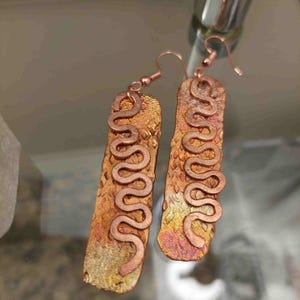 May include: A pair of copper earrings. Each earring features a rectangular, textured base with a copper serpentine design. The base has a mottled, warm color palette of gold, orange, and pink.