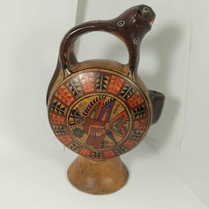 May include: A hand-painted, brown ceramic vessel with a circular design featuring a bird motif in red, orange, yellow, and green. The vessel has a handle and a spout, and is supported by a rounded base. The top of the vessel has a brown animal head.