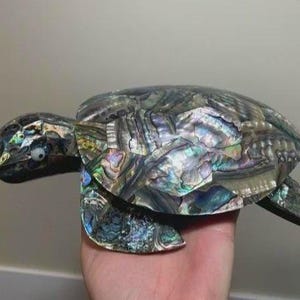 May include: A sea turtle figurine crafted from iridescent abalone shell pieces. The shell features a mosaic of shimmering green, blue, purple, and silver tones. The head and flippers are also covered in small, reflective shell fragments with a white eye.