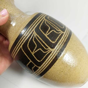 以下が含まれることがあります： 中央に濃い茶色の幾何学模様が施された、ベージュ色のセラミック花瓶。花瓶は細い首と丸みを帯びたボディをしています。デザインは線と形で構成されています。花瓶は人が持っています。