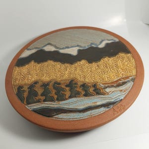 Op de afbeelding: Een ronde keramische schaal met een landschapsontwerp. De schaal heeft een gestructureerd, verhoogd ontwerp van een bos, bergen en een rivier in tinten bruin, goud, blauw en grijs. De rand is roodbruin.