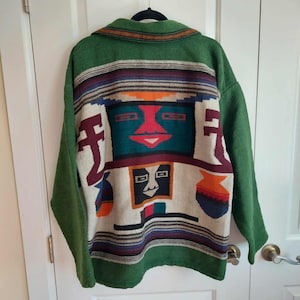 May include: A long-sleeved green jacket featuring a woven back panel with a multi-coloured geometric pattern. The design includes stylised face motifs in teal, red, and black, accented by horizontal stripes in orange, purple, and white.