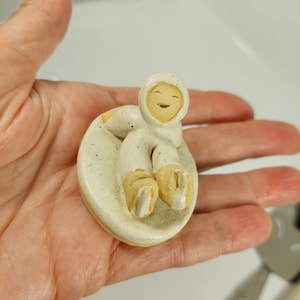 May include: A small ceramic figurine of a person in a hooded parka, seated on a round base. The figure is primarily off-white with speckled detailing, and the boots and face are a light tan colour. The figure has a smiling expression.