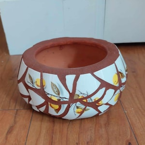 May include: A round ceramic planter with a mosaic design. The planter features a brown rim and interior, with a white exterior broken into irregular shapes. Yellow lemons and green leaves decorate the white surface.