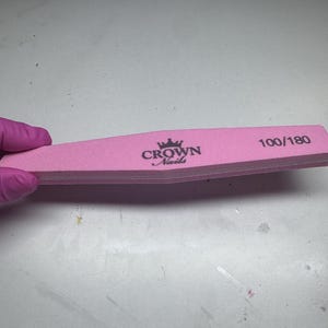 Peut inclure: Une lime à ongles rose avec le texte "CROWN Nails" et "100/180" imprimé dessus. La lime est tenue par une main gantée. La lime est de couleur rose clair.