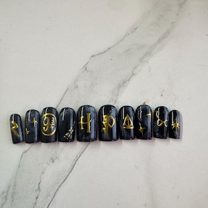 Puede incluir: Diez uñas postizas negras con purpurina dorada y símbolos de Harry Potter, incluyendo el escudo de Hogwarts, el símbolo de las Reliquias de la Muerte y el letrero de la Plataforma 9 3/4.