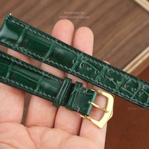 Puede incluir: Un par de correas de reloj verde esmeralda con un patrón de piel de cocodrilo. Las correas tienen hebillas doradas y se sostienen en una mano. El texto "Handmade by Vitalis Leather Goods" es visible.